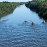 Cuatro pequeñas embarcaciones se desplazan por el Río Queguay con varias personas remando, a bordo. A los costados del Río, el eseso monte nativo llega hasta el agua
