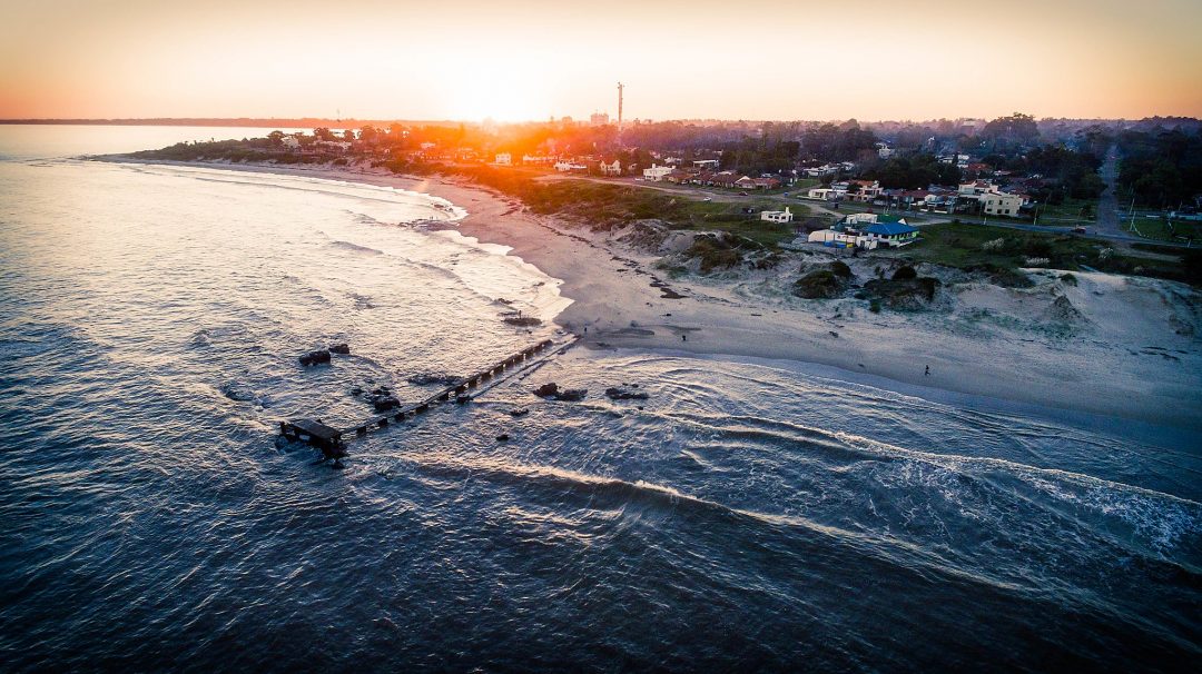 Vista aérea de un atardecer sobre Atlántida. Se observa el Río de la Plata y parte de la urbanización. Un muelle se adentra al mar