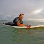 Juancho Posadas con traje de neopreno negro acostado sobre una tabla de surf blanca en el mar calmo