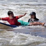 Un niño con camiseta roja extiende los brazos, sonríe y saca la lengua mientras una instructora detrás empuja su tabla para que barrene una ola