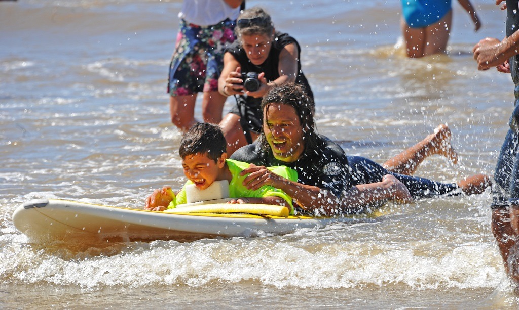 Un niño con una gran sonrisa se desliza acostado sobre una tabla de surf asistido por un guía ubicado detrás de él