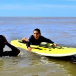 Niño sonrié sobre una tabla de surf amarilla con adaptaciones. A su costado, en el agua, un instructor de espaldas a la cámara se seca la cara