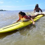 Una niña con camiseta blanca y roja sonríe y cierra los ojos mientras dos instructores detrás de ella empujan su tabla de surf cuando llega una ola