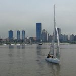 Un hombre navega en un velero adaptado de una plaza. Detrás se observa la silueta de la ciudad de Montevideo y el World Trade Center